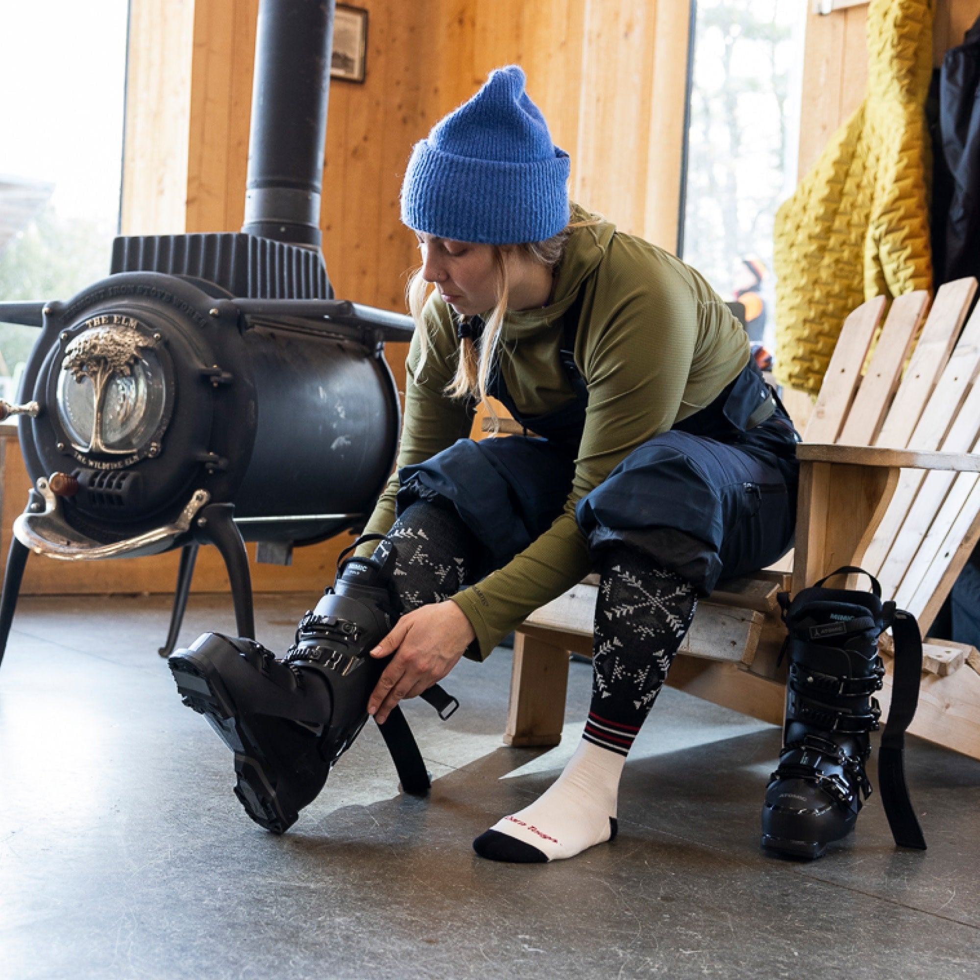 Model sitting on the edge of a chair next to a woodstove putting on ski boots wearing the Darn Tough Snowfall OTC sock in Black