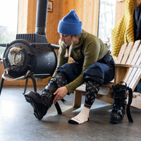 Model sitting on the edge of a chair next to a woodstove putting on ski boots wearing the Darn Tough Snowfall OTC sock in Black
