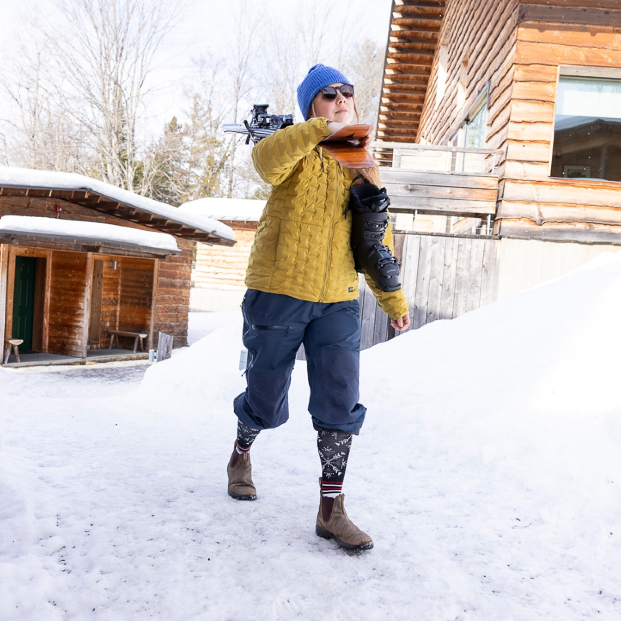 Model carrying skis and ski boots with snowpants rolled up showing the Darn Tough Snowfall OTC sock in Black